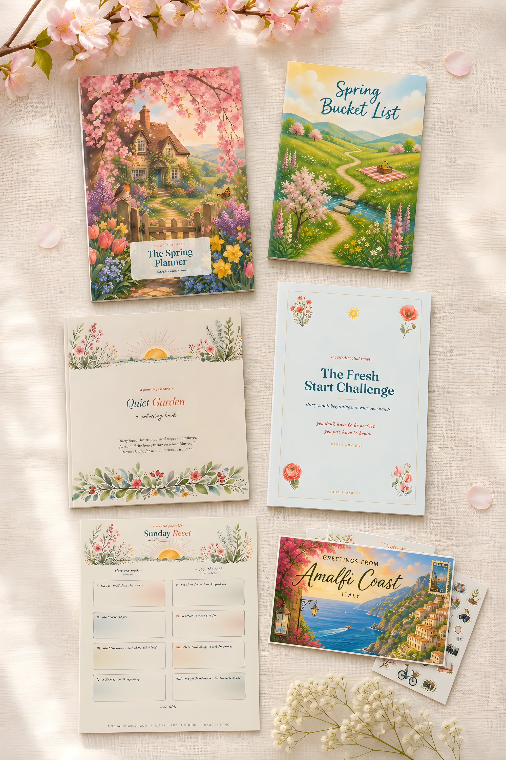 The Spring season's six pieces laid out on cream linen — planners, coloring book, reset sheet and postcards among blossoms